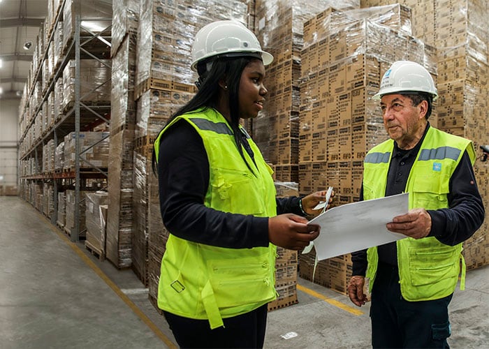Two workers wearing high?visibility vests and hard hats review documents together inside a warehouse with stacked pallets and shelving.