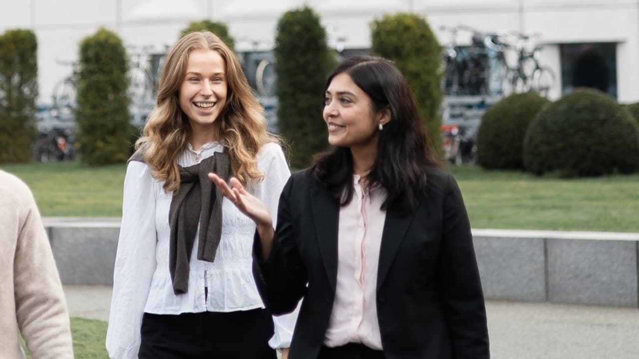 Two colleagues walking side by side outdoors, engaged in conversation, with office buildings, greenery, and bicycles in the background.