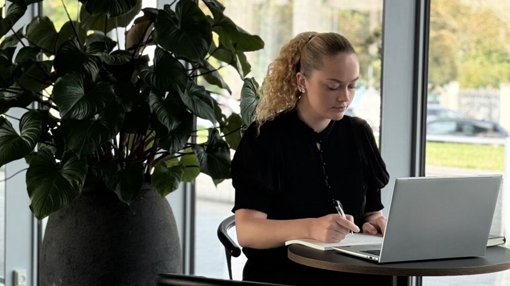 Person seated at a small table using a laptop and writing in a notebook, with a large indoor plant and office windows in the background.