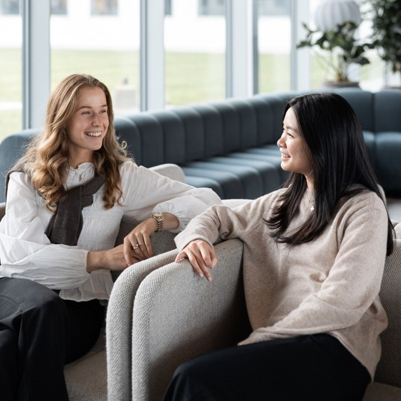 Two women sitting in a comfortable office lounge, smiling and engaged in a friendly conversation near large windows.