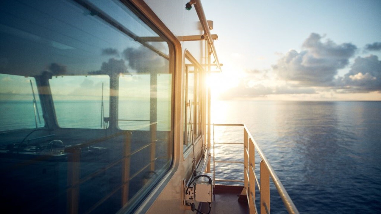View from a ship’s deck at sunrise, with sunlight reflecting off the ocean and clouds on the horizon, seen alongside the vessel’s windows and railing.