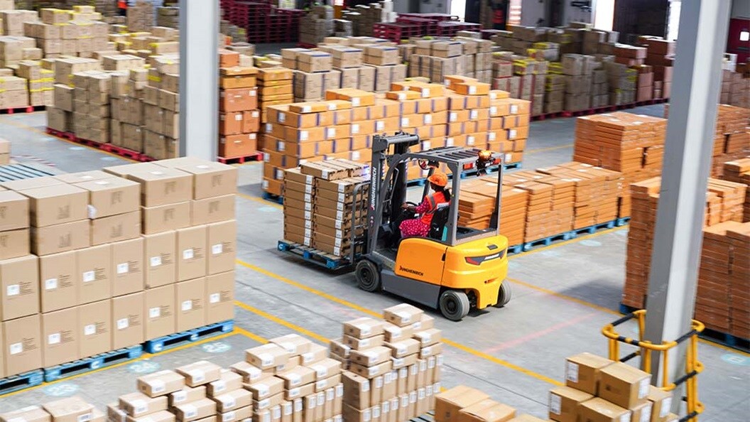 A worker drives a forklift through a warehouse filled with stacked boxes.