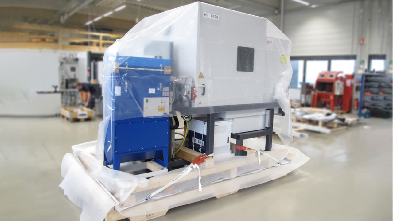 A machine covered in plastic is stored in a warehouse, indicating it is protected for transport or storage.