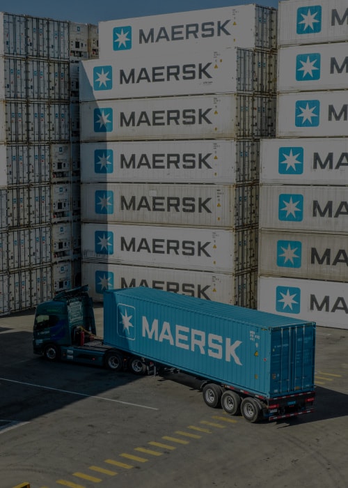 A container truck carrying a shipping container through a logistics terminal surrounded by stacked refrigerated and dry cargo containers